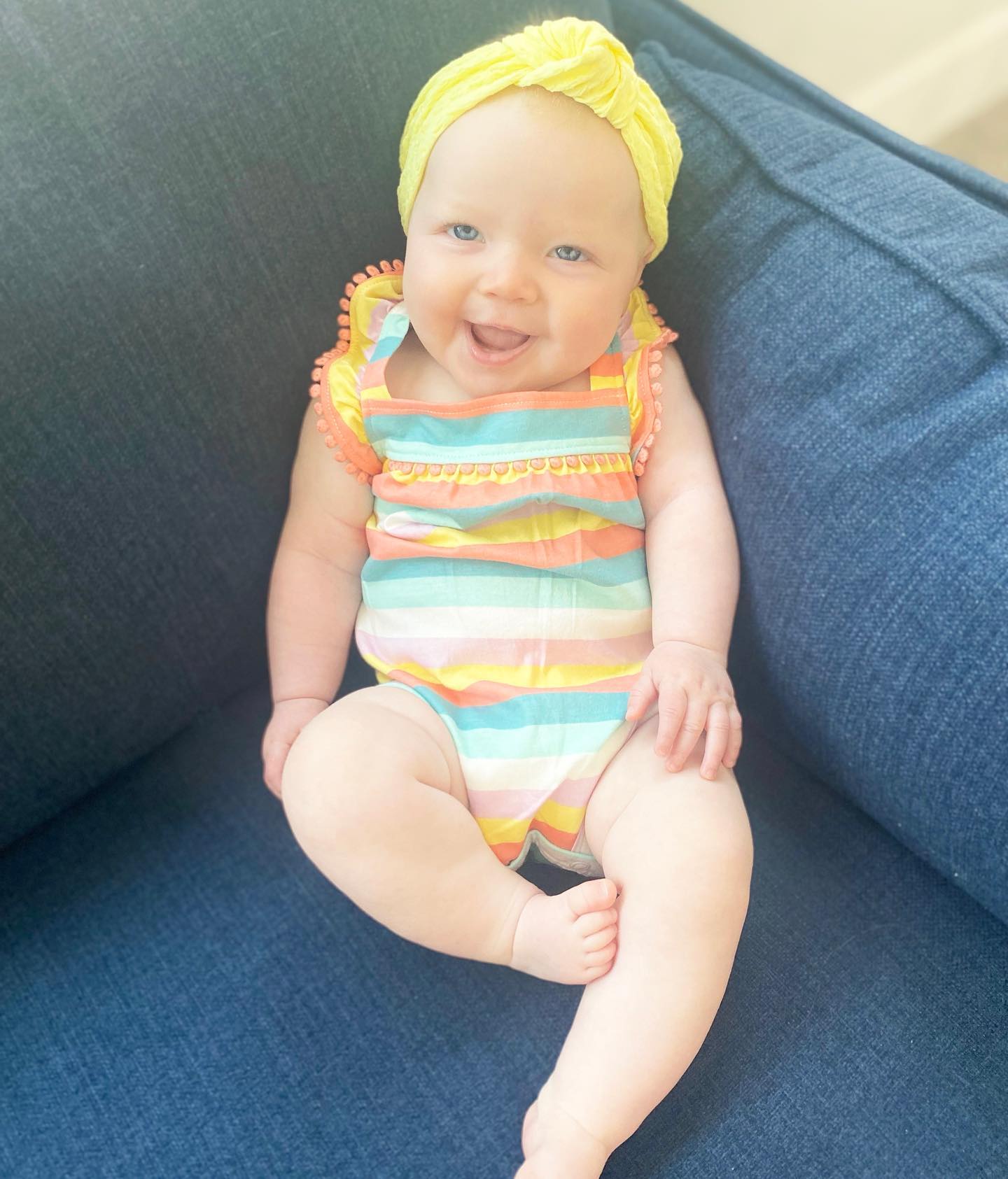 Happy baby in a striped romper and yellow headband smiling on a couch